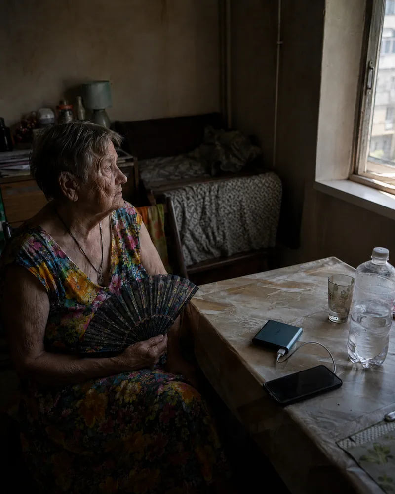 An older Ukrainian resident sitting beside a window with water, a phone, and a power bank during an outage.